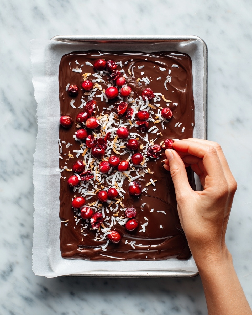 A rectangular layer of dark brown melted chocolate spread unevenly over white parchment paper on a metal tray, topped with scattered bright red dried cranberries that contrast against the glossy chocolate surface, and sprinkled with light beige coconut flakes adding texture; a woman's hand is seen mid-motion, gently placing more coconut flakes on the chocolate layer, against a white marbled background photo taken with an iphone --ar 4:5 --v 7