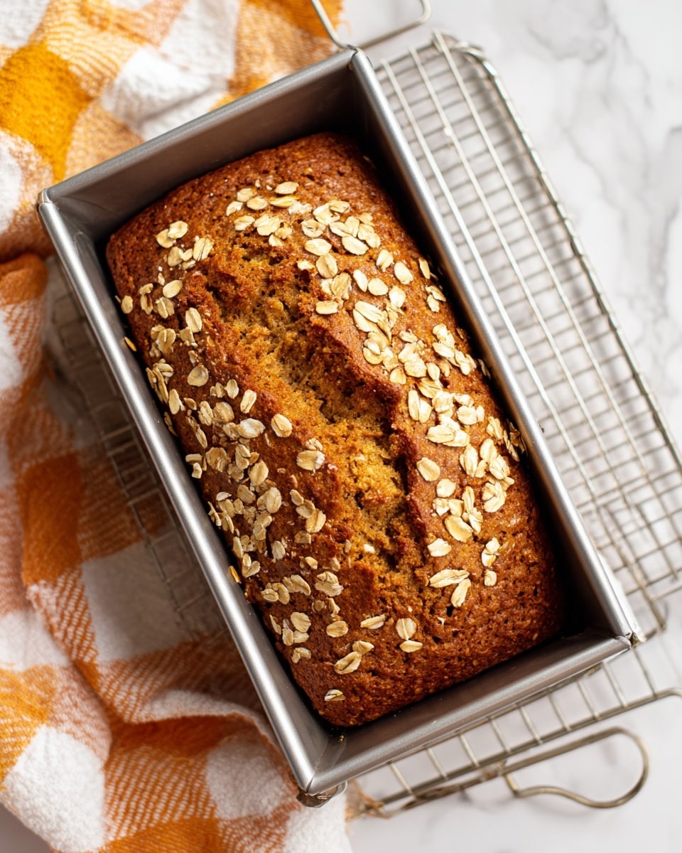 A loaf of bread in a silver metal rectangular baking pan topped with a thick layer of light beige oats scattered evenly across its golden brown crust. The crust has a slight crack running through the middle, revealing a soft, moist inside of warm brown color. The pan sits on a silver wire cooling rack over a white marbled surface, with a checkered cloth in white, orange, yellow, and black visible to the side. Photo taken with an iphone --ar 4:5 --v 7