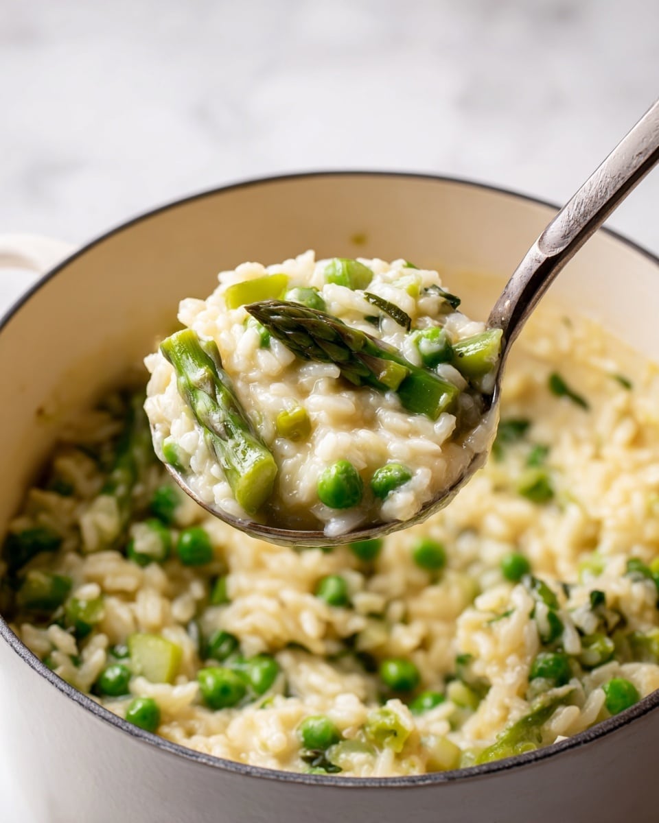 A close-up view of a white pot filled with a creamy rice dish mixed with bright green vegetables including peas and asparagus cut into pieces. The rice is light yellow and looks soft and slightly sticky, with a smooth texture. A metal spoon lifts a large scoop of the rice and vegetables above the pot, showing the mix clearly packed together. The background is a white marbled surface with a pale wall behind it. photo taken with an iphone --ar 4:5 --v 7