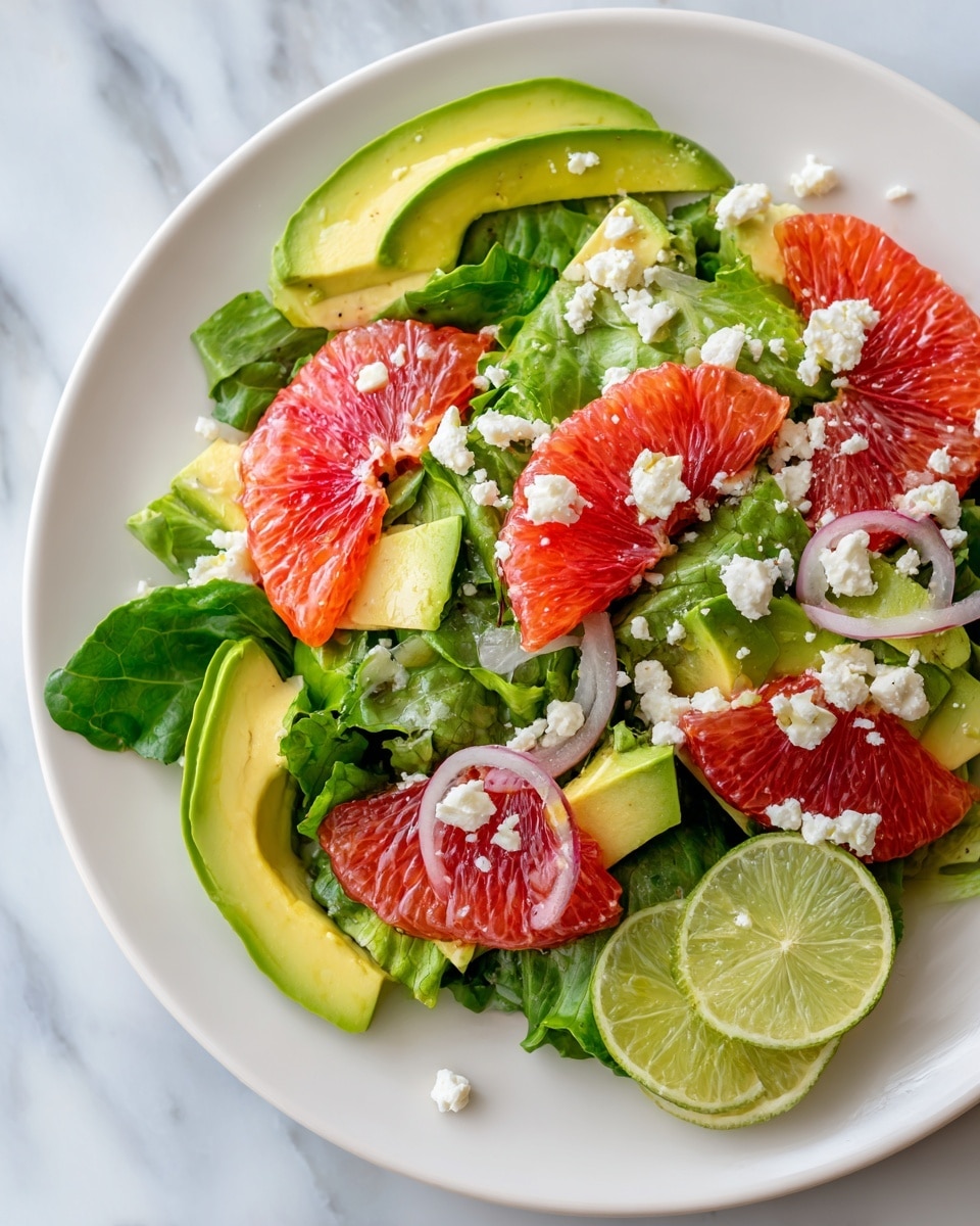 A fresh salad on a white plate with a mix of green lettuce leaves forming the base layer, topped with bright red blood orange pieces scattered evenly across. There are slices of creamy green avocado placed around the plate, mixed with small crumbles of white cheese sprinkled all over. Thin slices of light pink onion are visible among the greens, and two lime wedges are placed on the right edge of the plate. The plate sits on a white marbled surface. Photo taken with an iphone --ar 4:5 --v 7