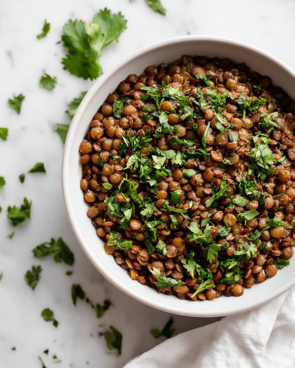 A close-up view of a bowl filled with cooked lentils that are brown and soft, topped with fresh green chopped cilantro leaves scattered across. The lentils appear moist and clumped but not mushy, creating a textured surface. The bowl is simple and white, placed on a white marbled surface with a bit of white cloth nearby. Some cilantro leaves are also lightly scattered on the surface around the bowl. Photo taken with an iphone --ar 4:5 --v 7