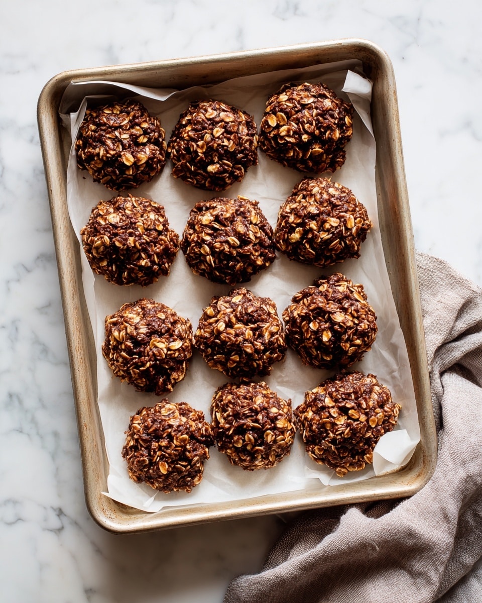 The image shows a metal baking tray lined with white paper holding twelve round, rough-textured chocolate and oat clusters arranged in three rows and four columns. Each cluster has a dark brown color with visible oats mixed in, giving a crunchy look. The tray is placed on a white marbled surface with a gray cloth napkin partially visible on the bottom right side. The clusters have an uneven, natural shape with small pieces sticking out, creating a textured surface. photo taken with an iphone --ar 4:5 --v 7