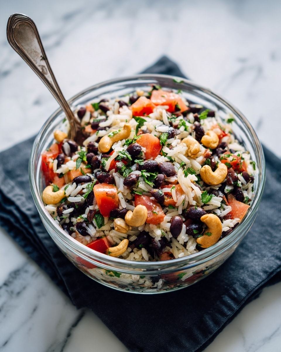 A clear glass bowl filled with a colorful salad placed on a dark cloth on a white marbled surface. The salad has layers of black beans, white rice, bright red tomato pieces, and light tan cashew nuts, all mixed together and topped with small green herb bits. A silver spoon rests inside the bowl on the left side. The overall look is fresh with a variety of textures and colors clearly visible. photo taken with an iphone --ar 4:5 --v 7