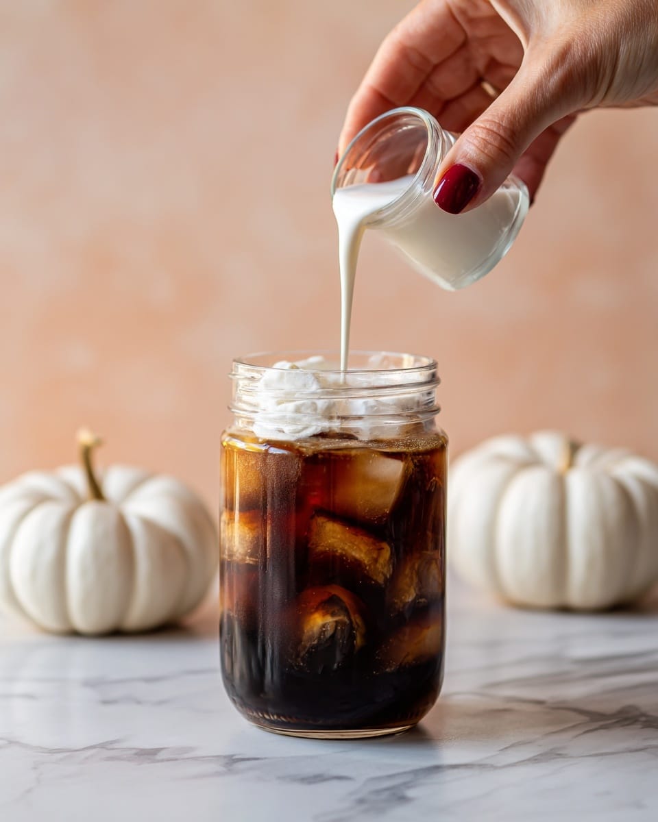 The image shows a clear glass jar filled with dark iced coffee and ice cubes. On top, a thick layer of white cream is being poured from a glass jar held by a woman's hand with dark red painted nails. The background has a soft peach color, and to each side of the jar are two white pumpkins. The jar sits on a white marbled surface. photo taken with an iphone --ar 4:5 --v 7