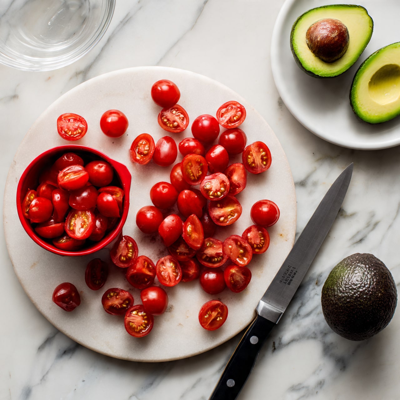 The image shows a round white marble board with a group of bright red cherry tomato halves scattered on it. A red measuring cup filled with more cherry tomato halves sits on the left side of the board. On the right side, there is a large knife with a black handle resting on the board next to the tomatoes. In the upper right corner, a white plate holds two halves of a ripe avocado, one with the large seed and the other seedless, along with a whole dark green avocado. A clear glass bowl is partially visible in the upper left corner on a white marbled surface. photo taken with an iphone --ar 4:5 --v 7