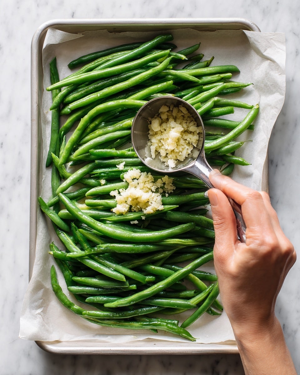 A white baking tray lined with parchment paper holds a single layer of fresh green beans spread out evenly. In the middle, there is a small pile of finely minced garlic adding texture and a pale yellow color. A woman's hand is holding a small metal measuring cup above the green beans, as if about to pour something. The background is a white marbled surface, bright and clean. photo taken with an iphone --ar 4:5 --v 7