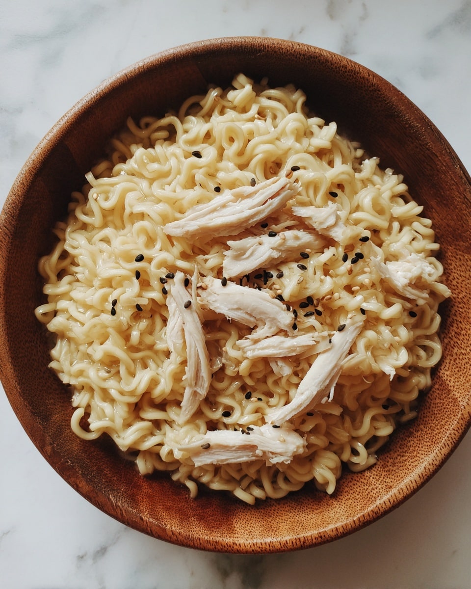 A wooden bowl filled with a single layer of cooked light yellow curly noodles topped with several pieces of pale white shredded chicken scattered evenly on top. Small black sesame seeds are sprinkled throughout the noodles and chicken, adding small dark spots for contrast. The bowl rests on a white marbled surface. photo taken with an iphone --ar 4:5 --v 7