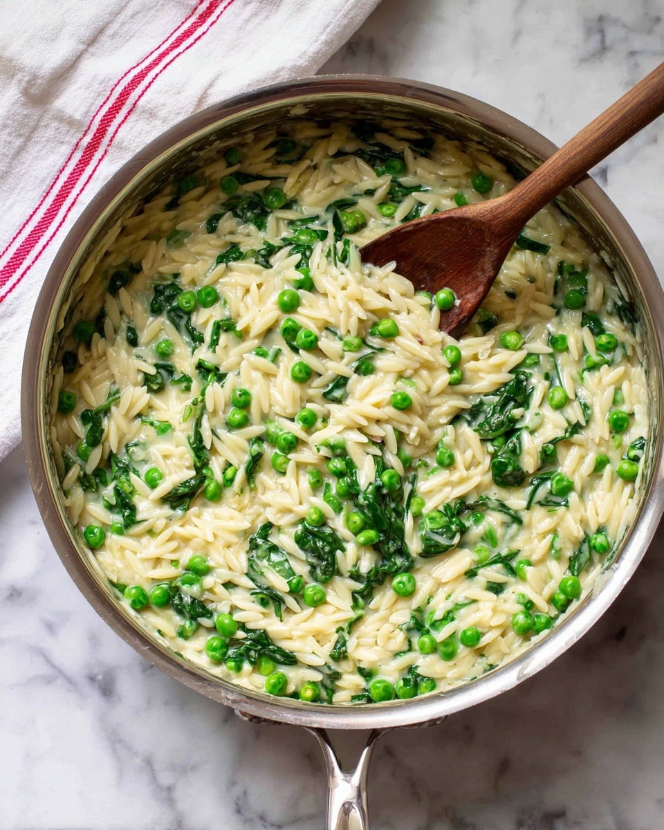 A white pan filled with orzo pasta mixed with bright green peas and dark green spinach leaves, all stirred with a wooden spoon positioned in the bottom right part of the pan. The orzo is light yellow with a soft, slightly glossy texture, while the peas are small and round with a fresh, smooth surface, and the spinach leaves are wilted but still vibrant. The pan is on a white marbled surface, and a white napkin with red stripes is folded partially on the left side of the image. photo taken with an iphone --ar 4:5 --v 7