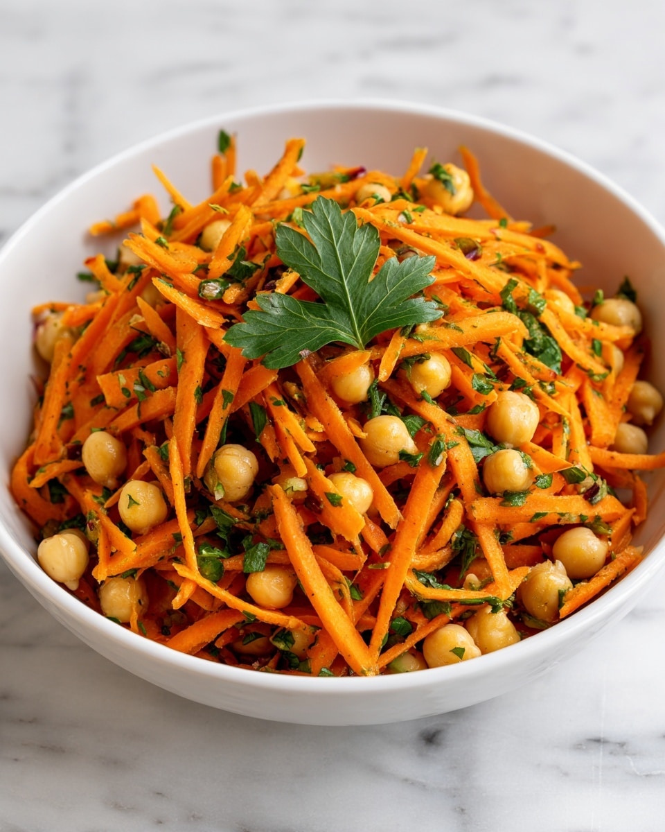 The image shows a white bowl filled with a salad made of thin orange carrot strips and light beige chickpeas mixed together. Bright green chopped herbs are scattered throughout the salad, with a few small whole green onion slices visible. On top, there is a small bunch of fresh green parsley leaves as garnish. The salad has a fresh, slightly glossy look, and it sits on a white marbled surface. photo taken with an iphone --ar 4:5 --v 7