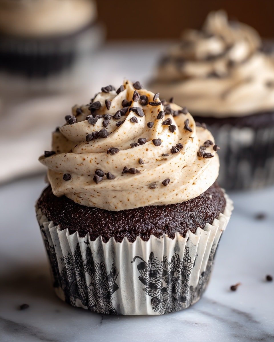 The image shows a close-up of a chocolate cupcake with a dark brown base and a white paper wrapper with black designs that is partially peeled away. On top of the cupcake, there is a thick layer of light beige frosting mixed with chocolate chips, giving it a textured look with small soft peaks. More chocolate chips are sprinkled on the frosting adding small dark brown spots. The cupcake sits on a white marbled surface with a blurred background showing another cupcake and a yellow bottle. Photo taken with an iphone --ar 4:5 --v 7