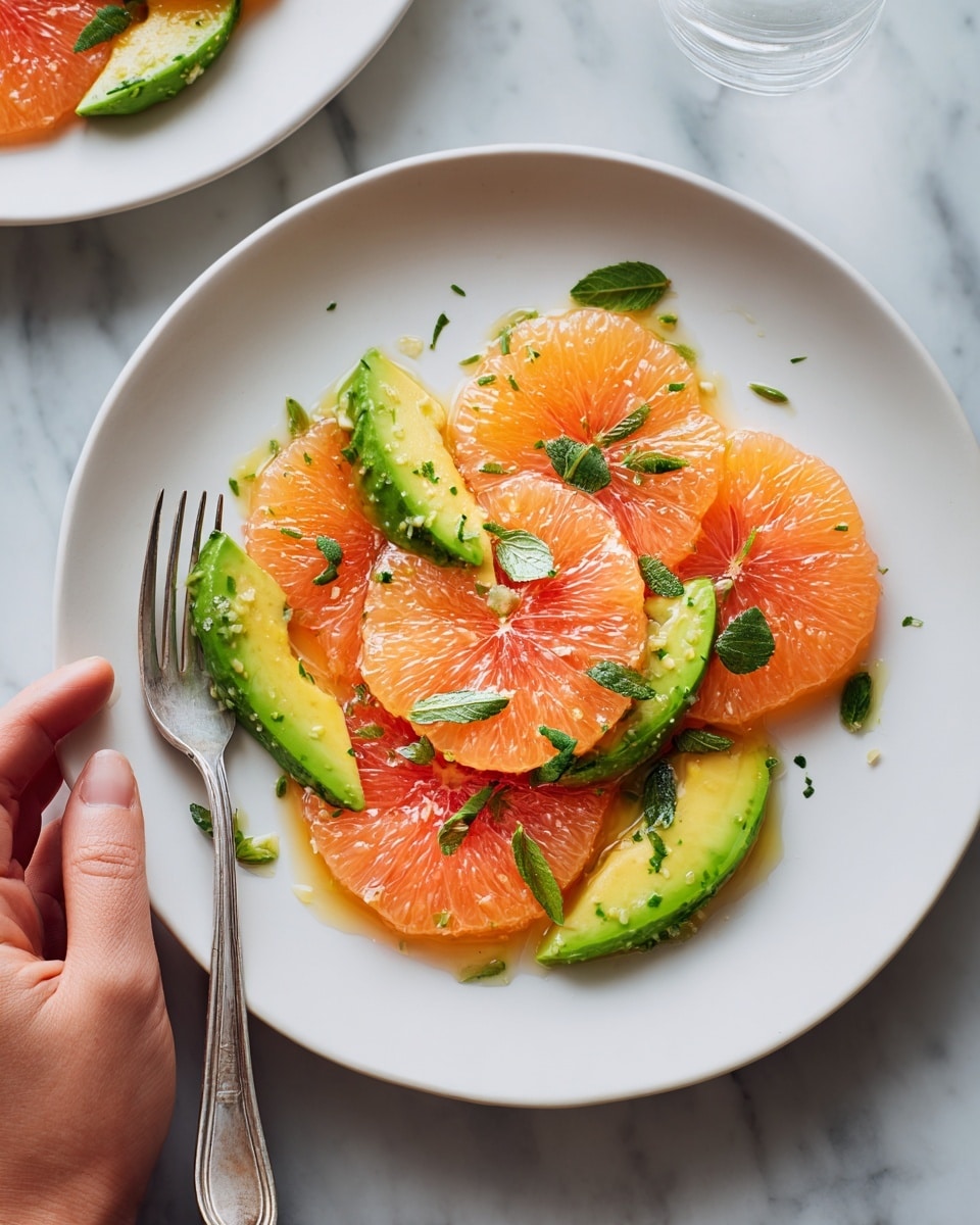 The image shows a white plate with thin slices of orange grapefruit and bright green avocado arranged in a circle. The grapefruit segments have a juicy, slightly shiny texture, while the avocado slices are smooth and creamy looking. Thin green herb leaves are sprinkled on top, adding small dark green spots for contrast. A silver fork rests on the left side of the plate, with a woman’s hand holding it gently. In the background, there is another white plate with the same salad partially in view. The whole setup is on a white marbled surface. photo taken with an iphone --ar 4:5 --v 7