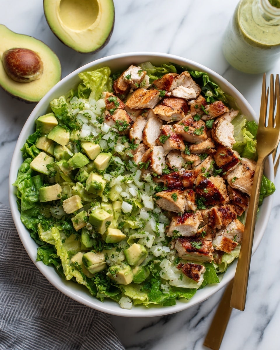 A white bowl filled with a green pasta salad made of spiral pasta mixed with chopped green herbs and small pieces of grilled chicken. The colors include light green pasta, dark green herbs, and browned chicken pieces. Two golden forks are placed in the bowl, with a white marbled surface under the bowl. Nearby are two avocado halves with green flesh and dark skin, and a small glass jar of green liquid dressing beside the bowl. A soft light beige cloth is on the upper left corner. Photo taken with an iphone --ar 4:5 --v 7