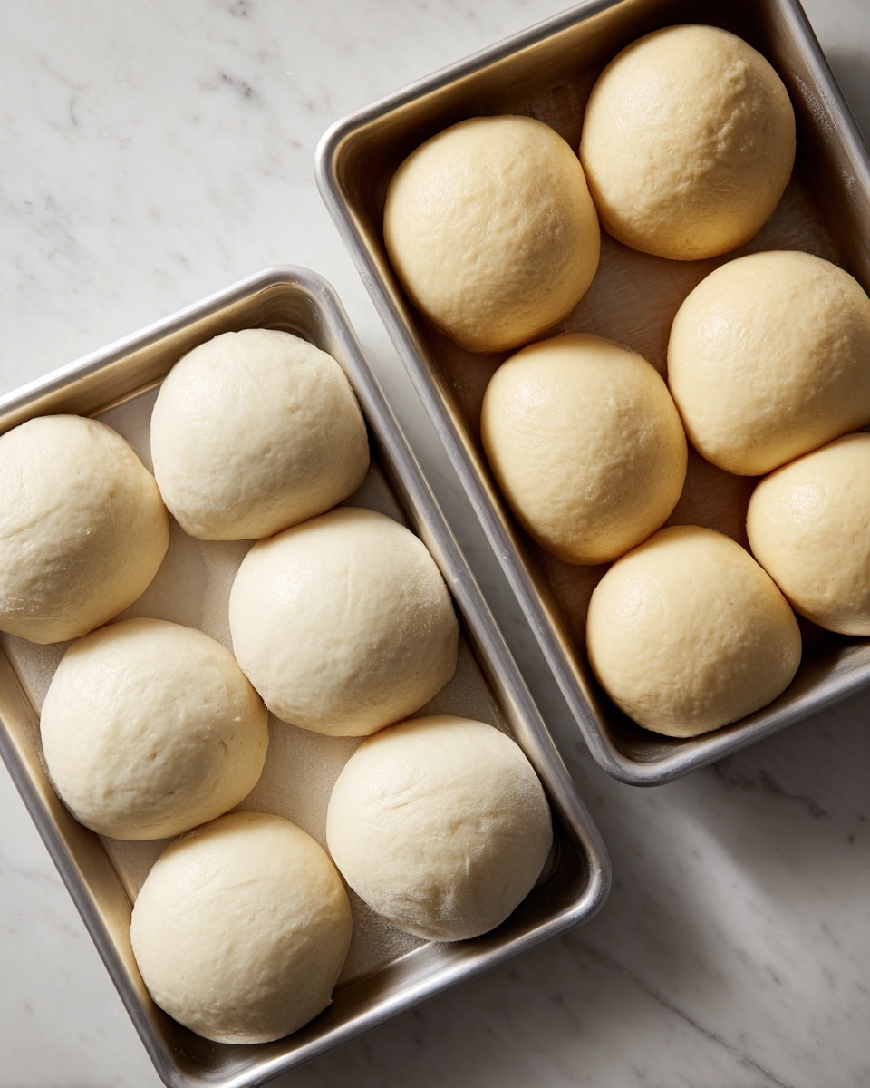 The image shows two rectangular silver trays placed on a white marbled surface. The tray on the left has four large pieces of dough, smooth and pale with a soft texture, evenly spaced in a single layer. The tray on the right holds ten smaller, round dough balls, light beige in color with a slightly shiny surface, tightly packed in a single layer filling the tray. The lighting highlights the smoothness and slight puffiness of the dough pieces and balls, making them look soft and ready for baking. Photo taken with an iphone --ar 4:5 --v 7