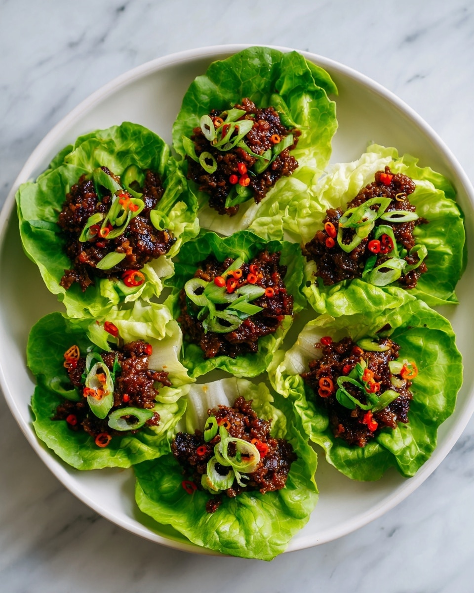 The image shows six fresh green lettuce leaves arranged on a large white plate with a white marbled surface underneath. Each leaf holds a generous portion of cooked ground meat mixed with small chopped vegetables and seasonings, all dark brown with hints of red and green from chili flakes and sliced green onions. The texture of the meat mix looks soft and crumbly, slightly glossy from sauce. Thin rings of fresh green onion are scattered over the meat on multiple leaves, adding a bright green contrast. The edges of the lettuce leaves are ruffled and fresh-looking, creating a layered and appetizing presentation. photo taken with an iphone --ar 4:5 --v 7