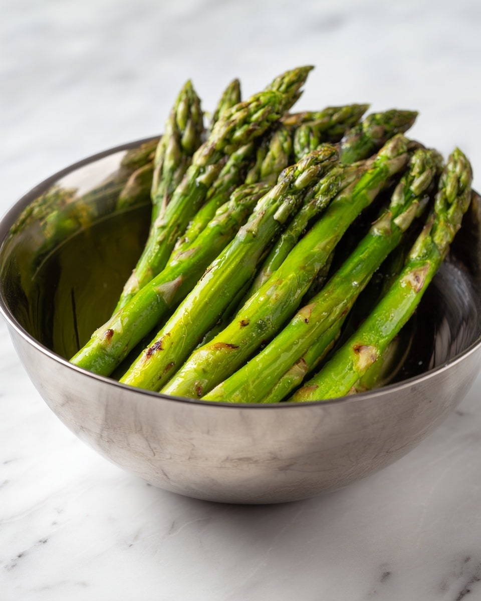 The image shows a shiny metal bowl on a white marbled surface filled with a bunch of green asparagus stalks. The asparagus are piled in the bowl, with bright green spears and some light brown grilled spots on the tips and sides. The metal bowl reflects light, creating smooth, shiny textures around the vegetables. Photo taken with an iphone --ar 4:5 --v 7