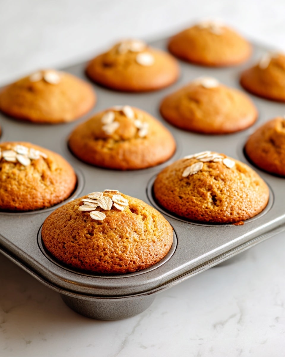 A metal muffin tray holds twelve freshly baked muffins, each one golden brown with a smooth top that rises slightly above the edge of the tray's round cups. Each muffin has a few scattered oats on top, adding a light cream color and rough texture against the soft golden surface. The tray sits on a white marbled surface, and the muffins show their soft, even texture with no cracks or unevenness. The light reflects softly on the slightly rough metal of the tray and highlights the gentle dome shape of each muffin top. photo taken with an iphone --ar 4:5 --v 7