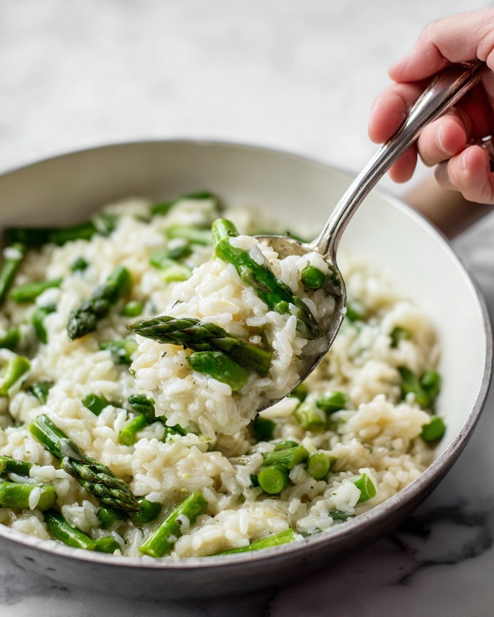 A close-up image of a white pan filled with cooked rice mixed with green asparagus pieces. The dish has a creamy texture, with rice grains looking soft and slightly sticky, and the asparagus adding bright green color and a slight crunch. A woman's hand is holding a spoon lifting a portion of the rice and asparagus mixture above the pan, showing the layers of rice and asparagus clearly. The background and surface are a white marbled texture. Photo taken with an iphone --ar 4:5 --v 7