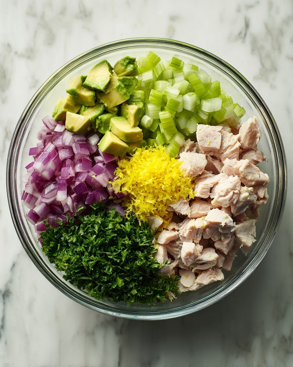 A clear glass bowl sits on a white marbled surface, filled with six distinct layers of ingredients arranged separately. Starting on the left side is a pile of green avocado chunks, slightly uneven in shape and size; next to it is a cluster of finely chopped pale green celery pieces. Moving upward, purple-red diced onion pieces are placed beside a small mound of bright yellow lemon zest. Above the lemon zest is a pile of chopped cooked chicken pieces in light pink and beige tones, with a soft, moist texture. Finally, dark green chopped parsley completes the circle on the upper left side, adding a fresh touch. The bowl holds all these layers clearly visible from above, ready to be mixed photo taken with an iphone --ar 4:5 --v 7