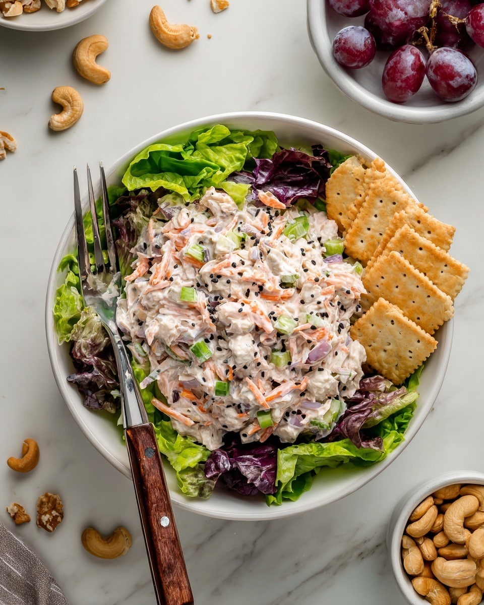 The image shows a white bowl filled with a layered salad. The bottom layer consists of fresh mixed green and purple leafy lettuce. On top, there is a thick layer of creamy chicken salad mixed with shredded carrots, small pieces of celery, and bits of red onion, all coated in a light-colored dressing with visible black pepper sprinkled on top. To the right side of the bowl, there are several light brown hexagonal crackers resting on the edge. A shiny silver fork with a wooden handle is placed inside the bowl on the left side, resting on the lettuce. The bowl is set on a white marbled surface scattered with a few cashew nuts and crumbs. Nearby, there are small white bowls filled with purple grapes and more cashew nuts. photo taken with an iphone --ar 4:5 --v 7