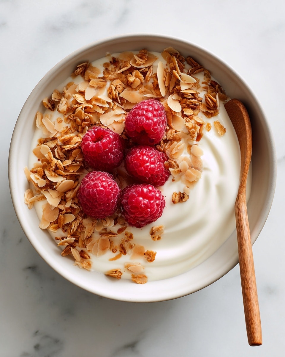 A white bowl filled with creamy white yogurt as the base layer, topped with clusters of golden brown granola mixed with oats and seeds spread evenly. Bright red raspberries are scattered on top, mostly in the center of the bowl, adding a fresh color contrast. A spoon with a wooden handle rests inside the bowl on the right side. The bowl sits on a white marbled surface with a light green cloth underneath. A woman’s hand holds the bowl from the top left corner photo taken with an iphone --ar 4:5 --v 7