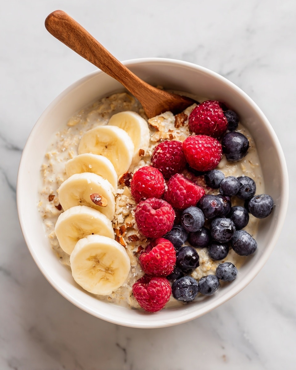 A white bowl filled with a creamy oatmeal base, topped with three layers of fruit: slices of light yellow banana arranged in a curved line near the edge, scattered bright red raspberries, and a handful of round, dark blue blueberries spread across the top. The oatmeal is mixed with visible small pieces of nuts and grains, and a wooden spoon rests inside the bowl on the left side. The bowl sits on a white marbled surface. photo taken with an iphone --ar 4:5 --v 7