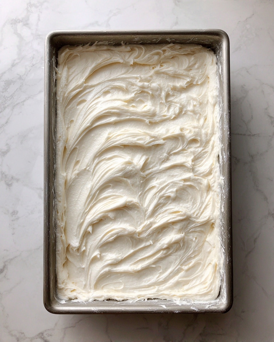 A rectangular metal tray filled with a smooth layer of creamy white frosting spread evenly, showing visible gentle swirls and peaks on the surface. The tray sits on a white marbled surface, with some frosting smudges on the tray edges, giving a fresh homemade look. photo taken with an iphone --ar 4:5 --v 7