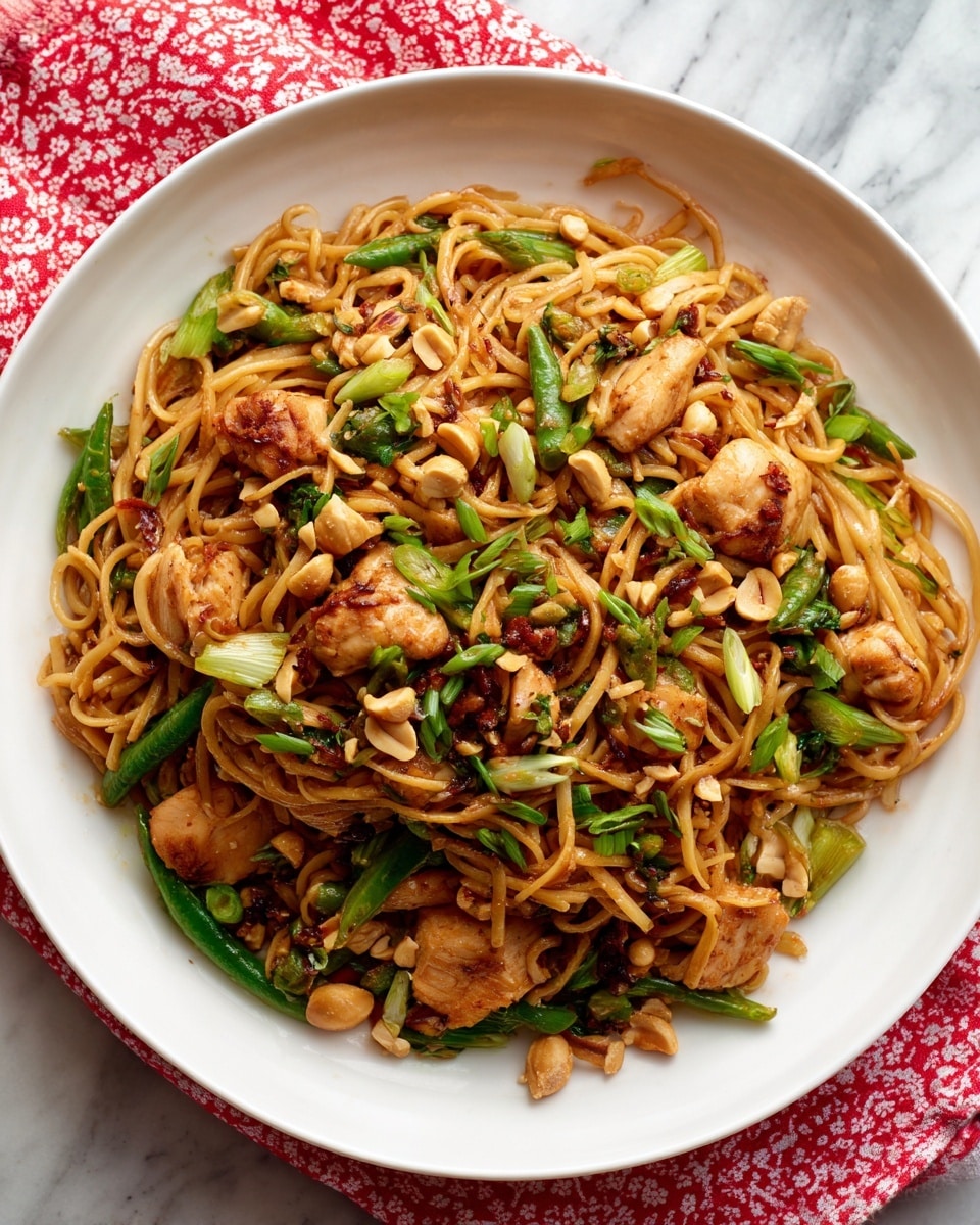 The image shows a white plate filled with a saucy noodle dish. The noodles are thick and orange-brown, mixed with chunks of meat that are darker and crispy on the edges. There are pieces of bright green vegetables scattered throughout, and the dish is topped with whole, light brown peanuts. The edges of the white plate have a red decorative pattern, and the plate sits on another white plate with the same red pattern. The whole setup is on a white marbled surface. The lighting highlights the glossy texture of the noodles and the peanuts, making them look fresh and tasty. Photo taken with an iphone --ar 4:5 --v 7