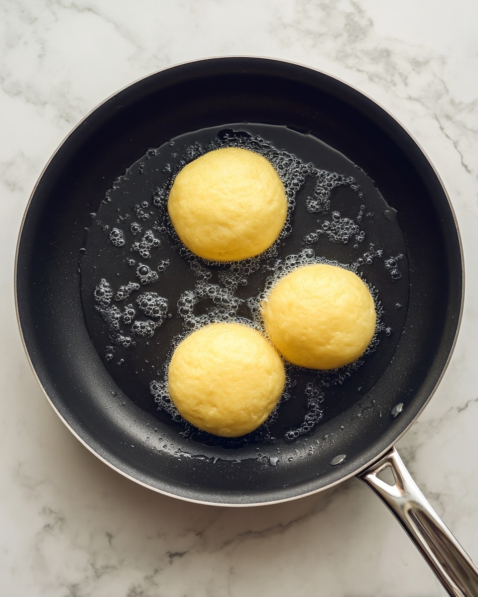 A close-up top view of three round dough pieces frying in a black non-stick pan with small bubbles of oil around the edges, placed on a white marbled surface. The dough pieces are light yellow with a smooth texture, evenly spaced in the pan, which has a shiny silver handle reflecting light. Photo taken with an iphone --ar 4:5 --v 7