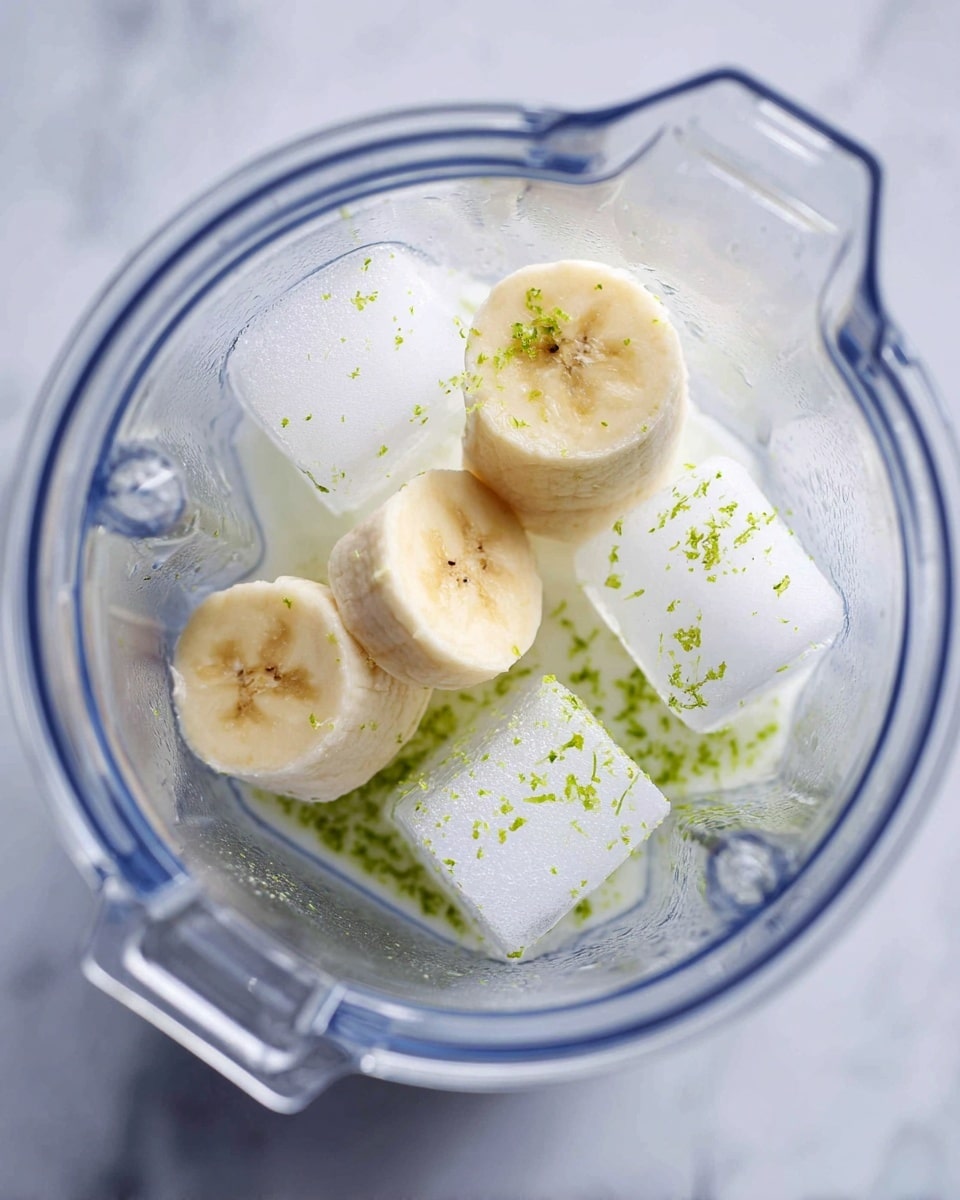 The image shows a clear blender container viewed from above, filled with four ice cubes scattered around the bottom, two thick slices of banana placed near the center, and a small amount of white liquid that almost covers the base. Tiny green lime zest bits are spread lightly on the surface and sides inside the blender. The blender is set on a white marbled surface. Photo taken with an iphone --ar 4:5 --v 7
