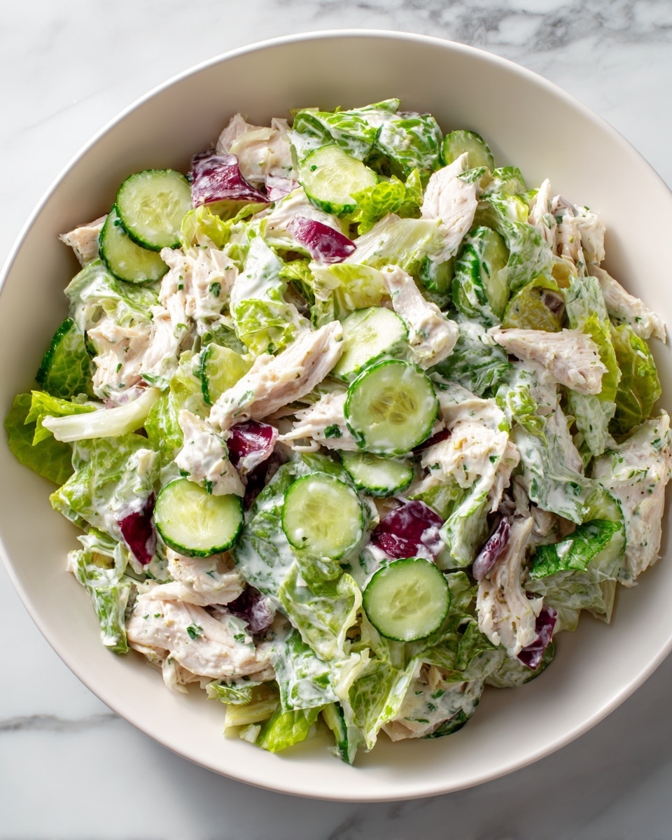 A close-up of a white bowl filled with a creamy salad showing shredded white chicken pieces mixed with bright green lettuce leaves, circular slices of cucumber with dark green skin, and chunks of purple-red onion. The salad is thickly coated in a white, creamy dressing that clings to all the ingredients, giving a moist texture. The bowl is placed on a white marbled surface, highlighting the fresh and colorful mix inside. photo taken with an iphone --ar 4:5 --v 7