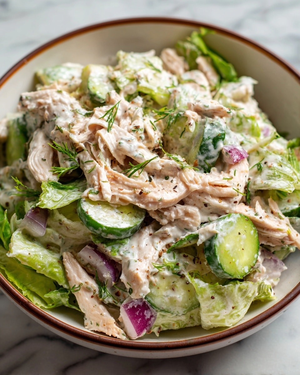 A white bowl filled with a creamy salad that has three main layers: shredded light beige chicken pieces mixed with green leafy lettuce and bright green cucumber slices, all coated in a white creamy dressing. There are chunks of purple onion scattered throughout, adding a splash of color. The bowl sits on a white marbled surface. Photo taken with an iphone --ar 4:5 --v 7