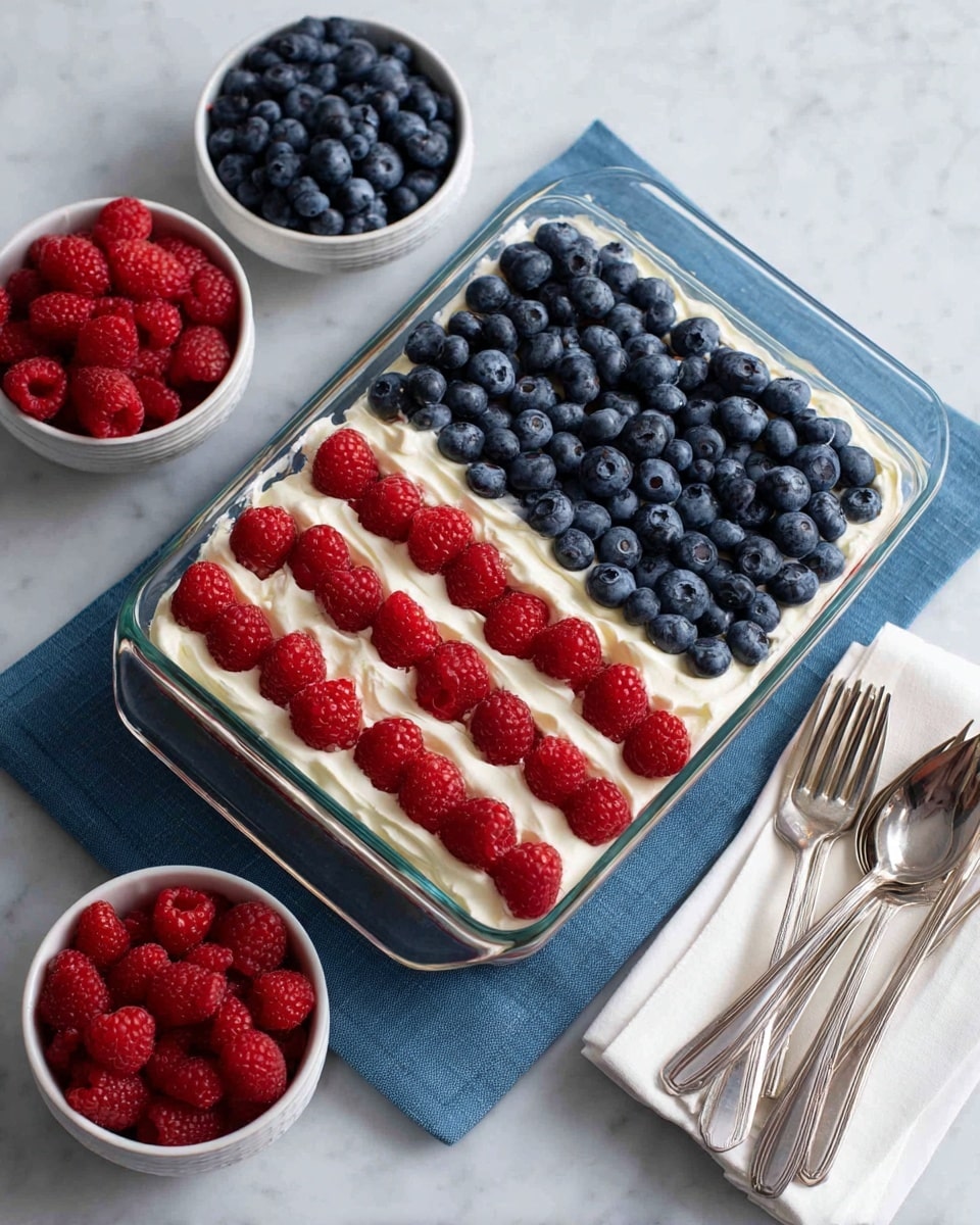 A rectangular glass dish filled with a dessert arranged to look like the American flag. The bottom layer is white cream or frosting. There are horizontal rows of alternating white cream and red raspberries, making seven red stripes and six white stripes. In the top left corner, a square area is covered with fresh blueberries. Around the dish are two small white bowls, one filled with blueberries and the other with raspberries, placed on a white marbled surface with a blue cloth beneath the dish. To the right, there is a set of white napkins with silver forks on top. Photo taken with an iphone --ar 4:5 --v 7