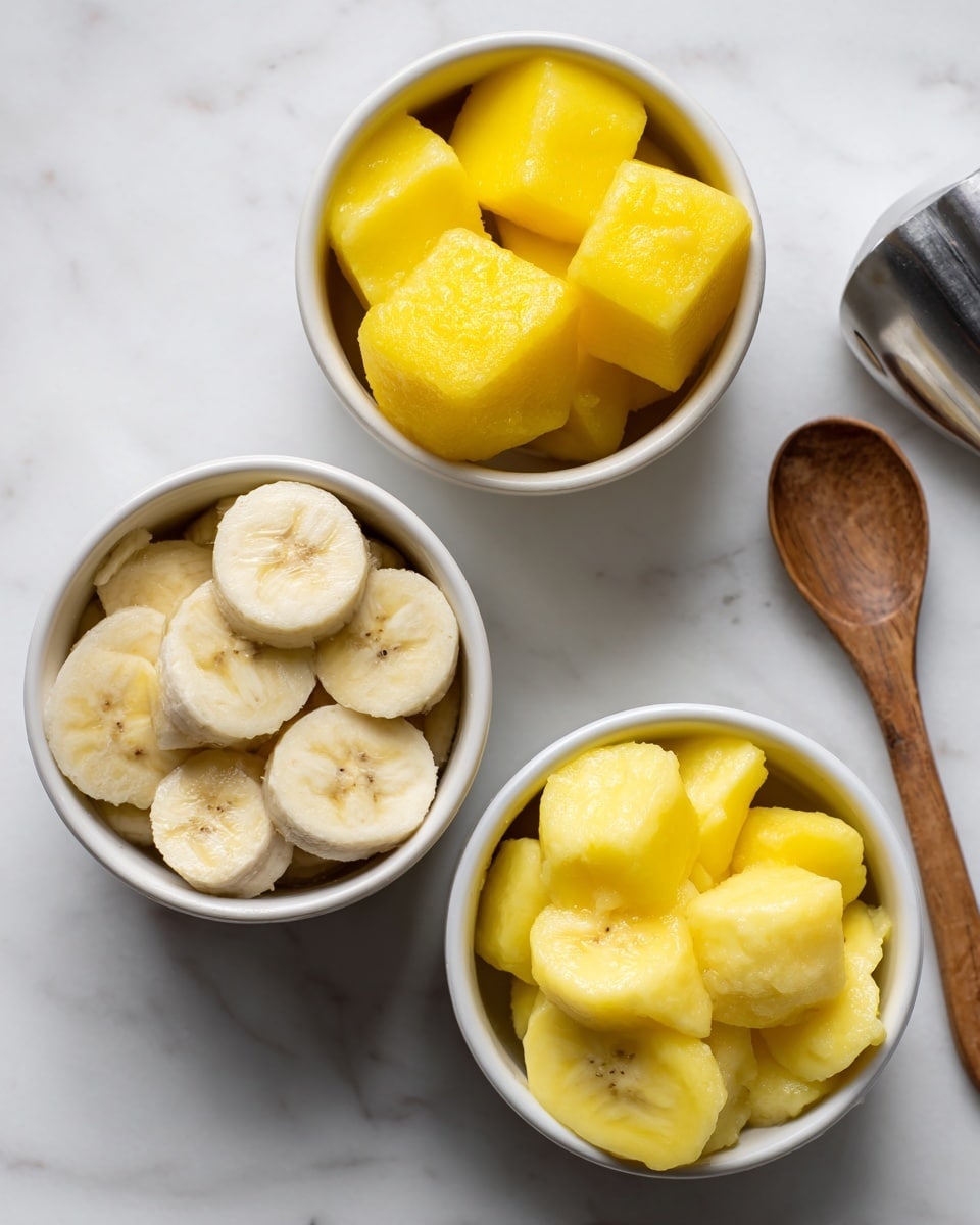 The image shows three small white bowls placed on a white marbled surface. The top left bowl holds bright yellow frozen mango chunks, with a smooth icy texture. The bottom left bowl contains circular, light beige banana slices, soft and fresh. The bottom right bowl is filled with more bright yellow frozen mango pieces similar to the first bowl. To the top right side, there is a small wooden spoon resting on the surface. A silver kitchen mixer is partly visible at the upper right edge. The overall scene is neat and bright, showing the fruits clearly. photo taken with an iphone --ar 4:5 --v 7
