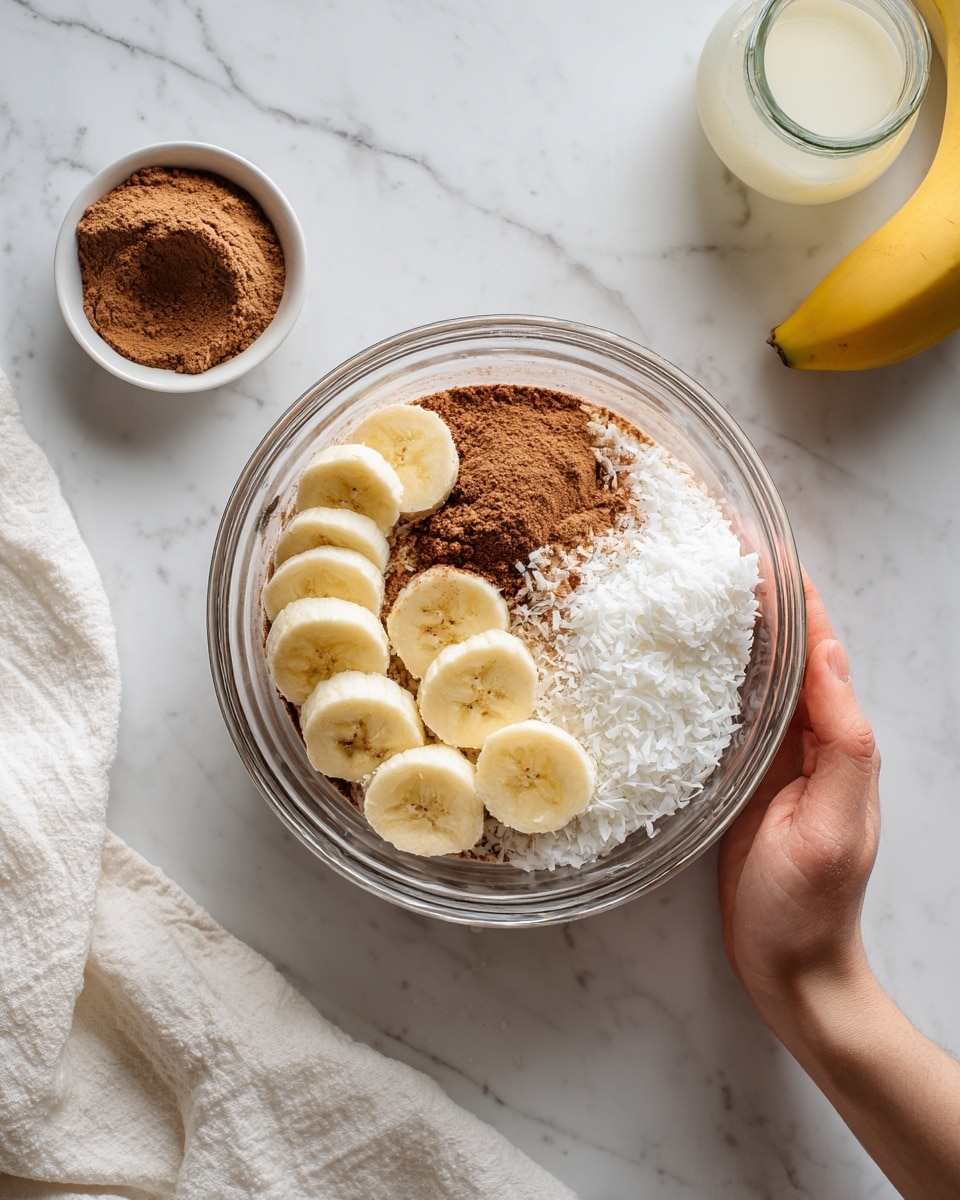 The image shows a clear glass mixing bowl on a white marbled surface, filled with several layers of ingredients. At the bottom, a layer of white coconut shreds is visible, topped with brown powder, likely cinnamon or cocoa. On one side inside the bowl, there are several thick banana slices arranged, light yellow with soft texture. Outside the bowl, to the left, is a small white bowl holding more brown powder, and to the right, a glass bottle with light-colored liquid. A peeled banana and a white cloth are visible in the background. A woman's hand is holding the bowl gently from the right side. photo taken with an iphone --ar 4:5 --v 7