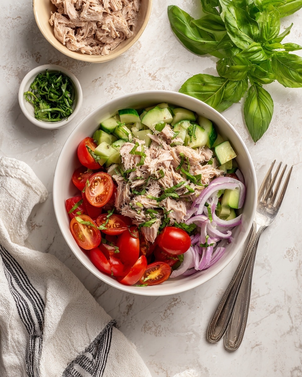 A large white bowl filled with a colorful salad showing three main layers: the base layer of bright red halved cherry tomatoes, fresh green chopped cucumber pieces, and torn leafy greens. Scattered on top, chunked light tan tuna pieces create the second layer, mixed with thin, semi-transparent purple onion slices and small green herbs. Two shiny silver forks rest inside the bowl on the right side. The bowl sits on a white marbled surface with a white and black striped cloth slightly underneath on the left side. Nearby, fresh green basil leaves and part of a brown bowl with extra tuna pieces are visible at the top and right sides. Photo taken with an iphone --ar 4:5 --v 7