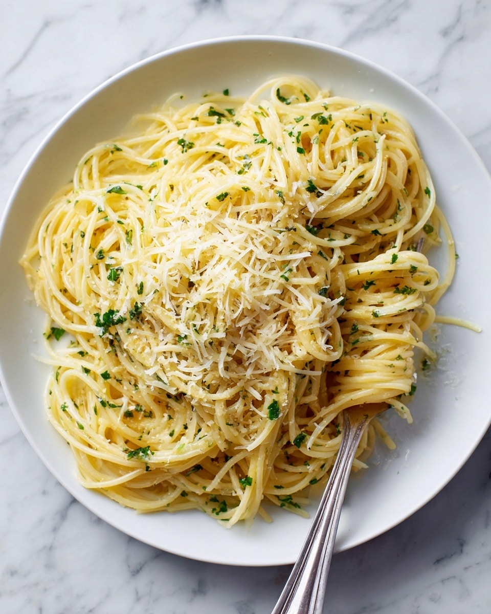 A white plate holds a serving of spaghetti pasta with a creamy light yellow sauce. The pasta is mixed with small green herb pieces scattered evenly throughout. On top, a layer of finely grated pale yellow cheese is sprinkled, adding texture. A silver fork rests on the plate, with some pasta wrapped around its tines, positioned near the right side. The plate sits on a white marbled surface. photo taken with an iphone --ar 4:5 --v 7