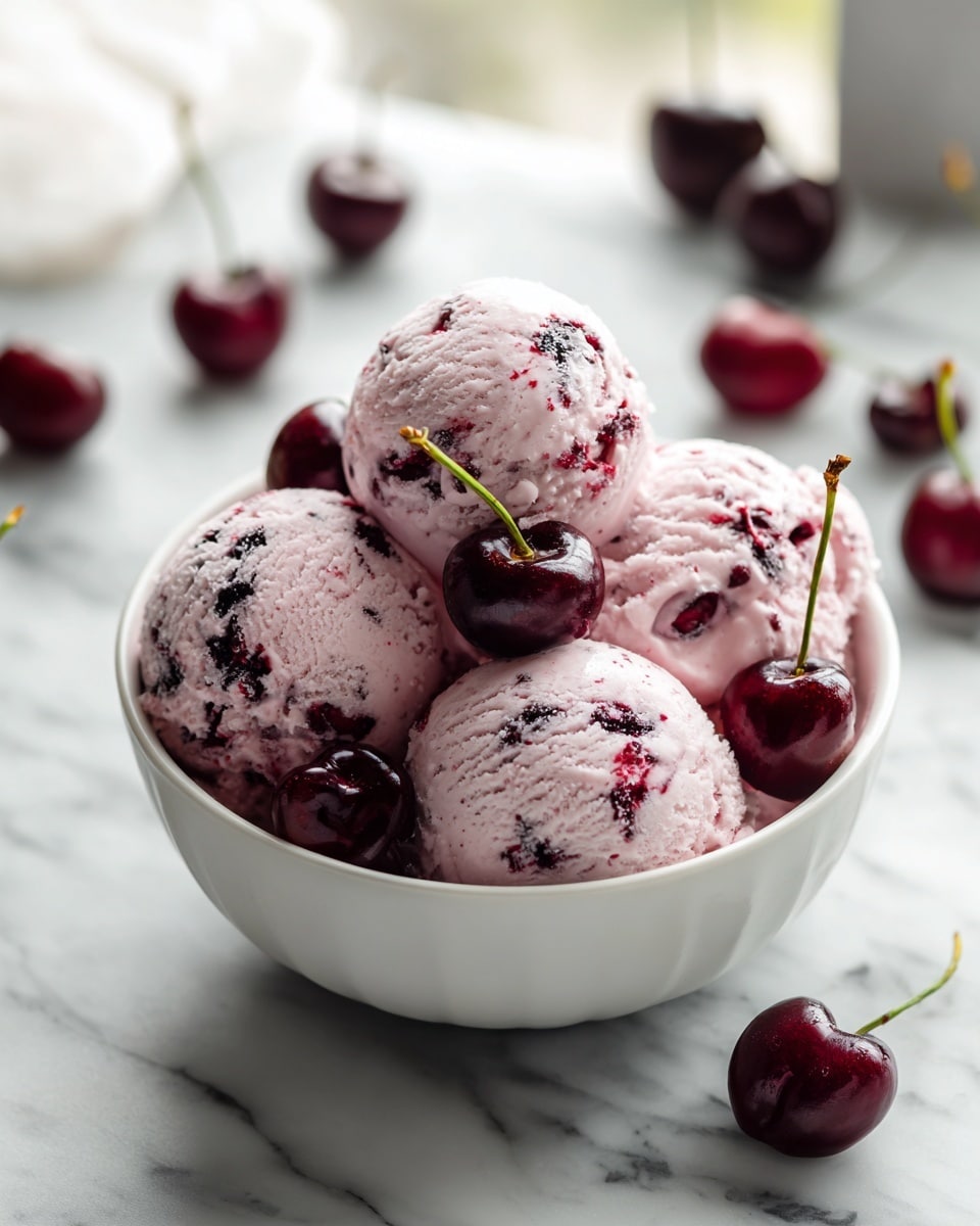 A white bowl filled with five scoops of light pink cherry ice cream, each scoop showing dark red cherry bits mixed within. Dark whole cherries are placed on top of the scoops and around the bowl on a white marbled surface. The bowl has a ribbed texture and the background shows a soft-focus kitchen. photo taken with an iphone --ar 4:5 --v 7