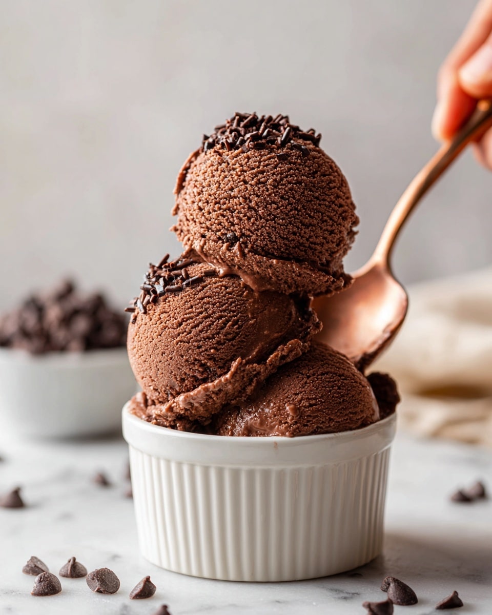 The image shows three scoops of rich, dark chocolate ice cream stacked in a small white ceramic bowl. The ice cream looks smooth and creamy, with a slightly shiny surface and small chocolate flakes sprinkled on top. Around the bowl are dark chocolate chips scattered on a white marbled surface. A woman's hand holds a bronze spoon next to the bowl, ready to dig in. The light makes the chocolate color look deep and inviting. photo taken with an iphone --ar 4:5 --v 7