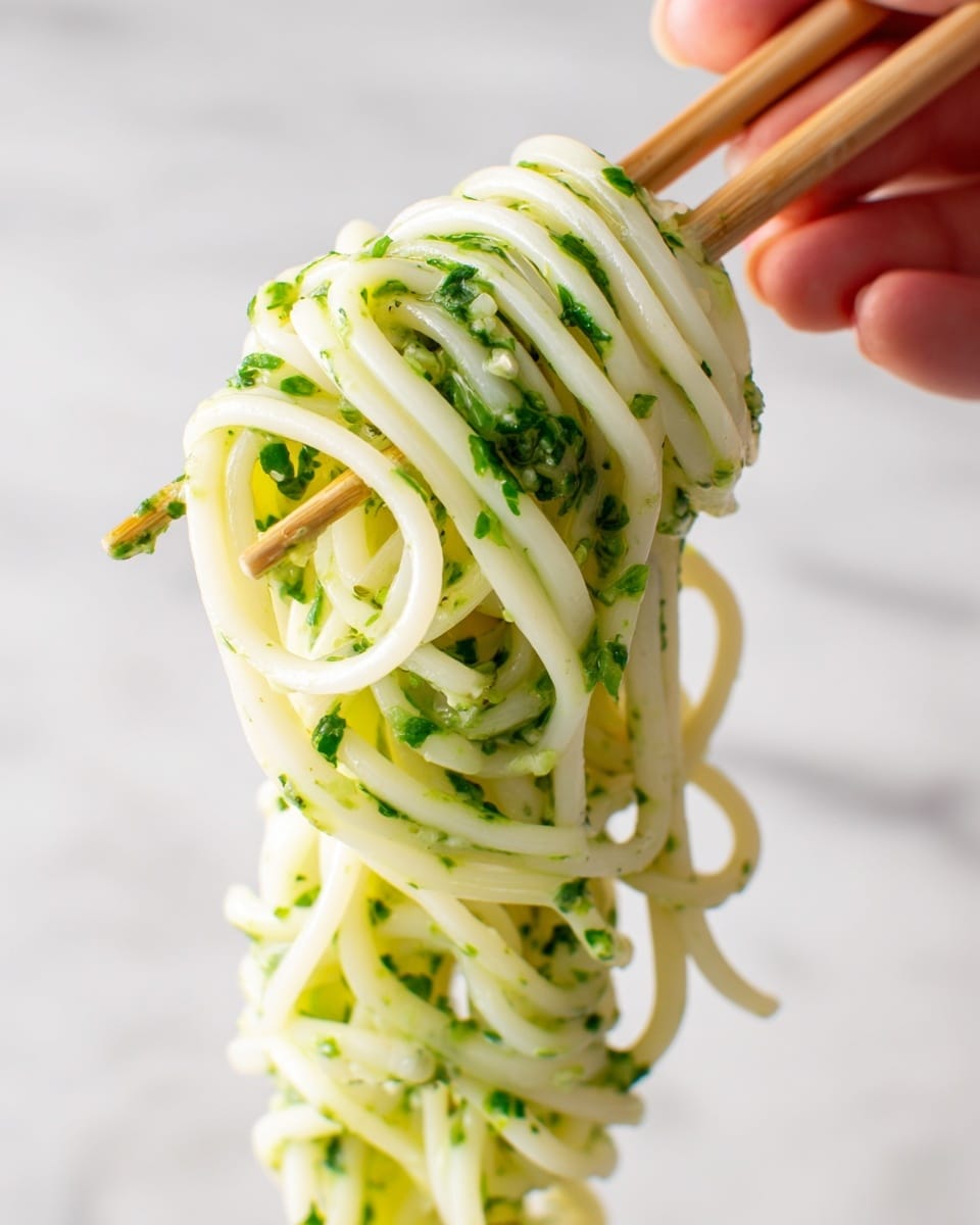 Thin white pasta noodles are lifted closely by a pair of dark metal tongs, showing a thick green sauce with small pieces of herbs spread unevenly on the noodles. The noodles have a smooth and shiny texture, hanging down in long strands with some parts showing the sauce clinging to them. The background is plain with a white marbled texture. Photo taken with an iphone --ar 4:5 --v 7