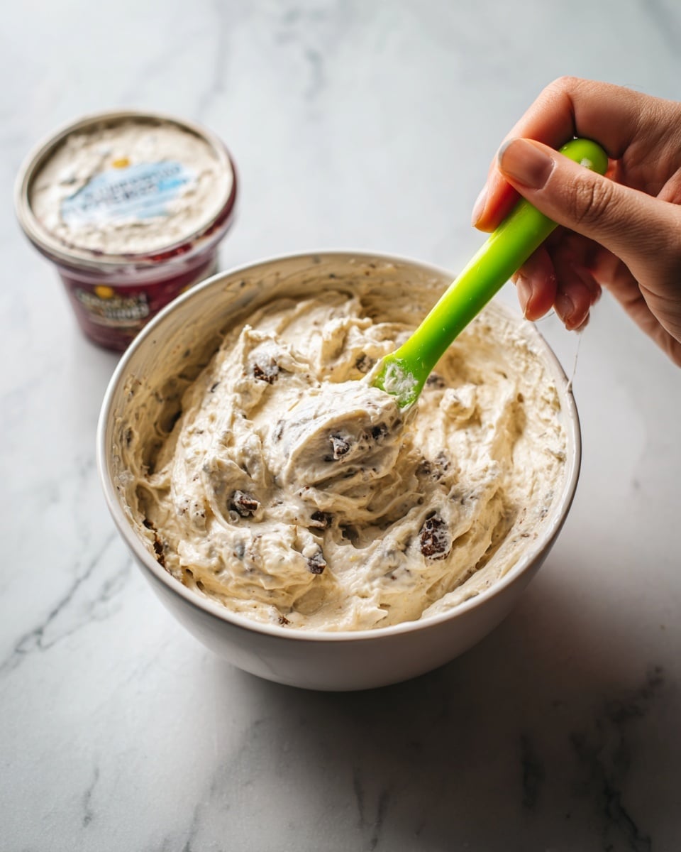 In the image, there is a white bowl filled with a creamy mixture that has a light beige color with dark bits scattered throughout. A woman's hand is holding a spoon with a bright green handle, stirring the thick, slightly lumpy texture inside the bowl. Next to the bowl is a round container of cottage cheese with a red and white label. The surface beneath everything is white with light marble veins. The lighting is soft and natural, making the texture of the mixture clear and inviting. Photo taken with an iphone --ar 4:5 --v 7