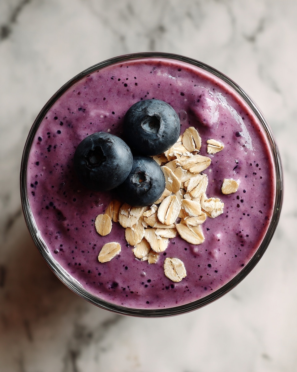 A close-up top view of a creamy purple smoothie in a clear glass. The smoothie is smooth with tiny dark specks throughout. On top, there are three fresh dark blue blueberries arranged in a triangle, surrounded by a few light tan rolled oats scattered around them. The glass sits on a white marbled surface with a soft blurred pink cloth in the background. Photo taken with an iphone --ar 4:5 --v 7